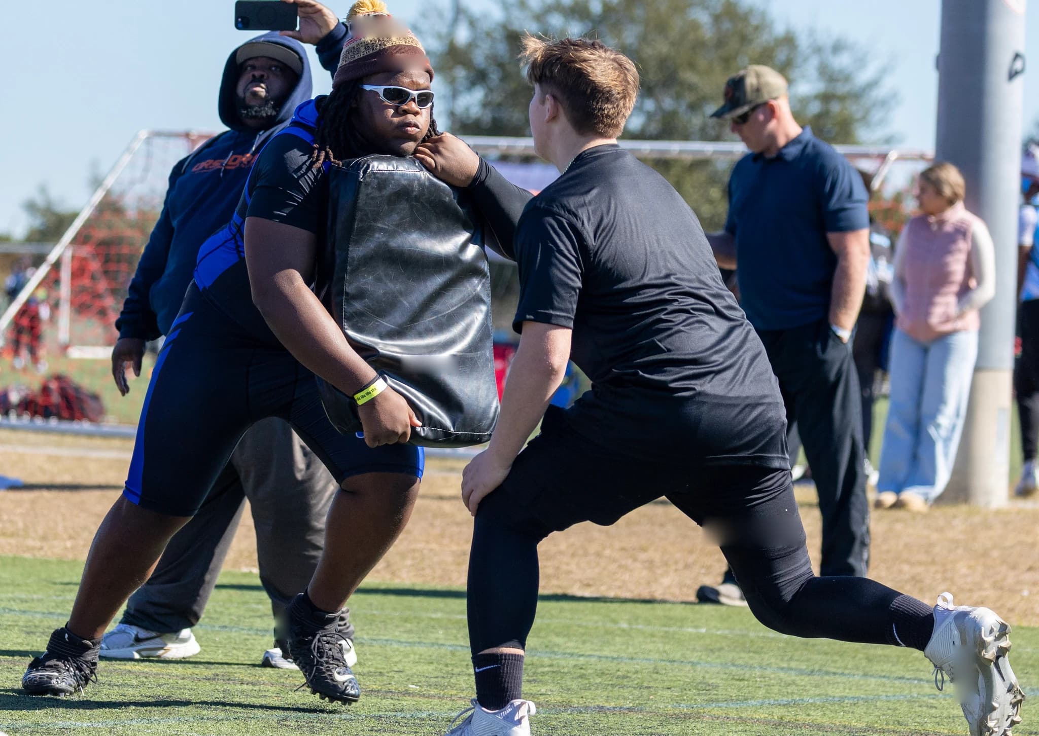 Linemen competing in a blocking drill with an adult observing and sideline activity being recorded